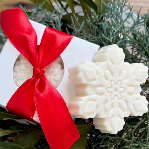 Decorative soap with snowflake design and red ribbon in a white box, surrounded by greenery.